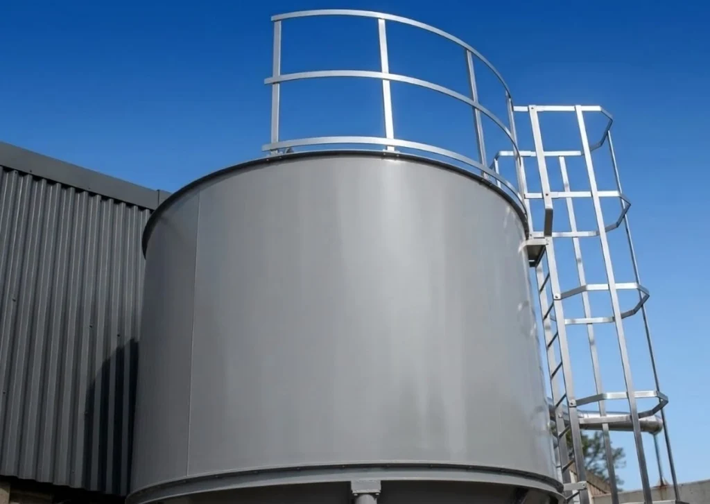 Cylindrical grey tank with a metal access ladder and guardrail at the top, next to a corrugated metal building under a blue sky