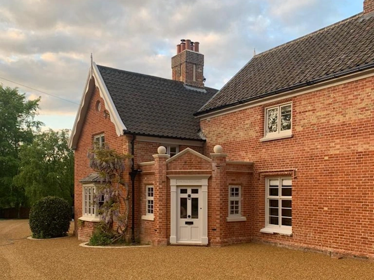 Restored red brick country house with a tiled roof, white-framed windows, a pedimented porch and a wisteria climbing the wall above a gravel driveway