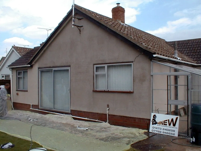 Rendered bungalow with sliding patio doors and sheeting laid on the ground, a worker in a protective suit at the left and a blast cleaning sign by the greenhouse
