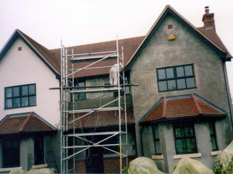 Two-storey house with grey and cream rendered walls and a tiled roof, with an aluminium scaffold tower and a worker in a white protective suit on the platform