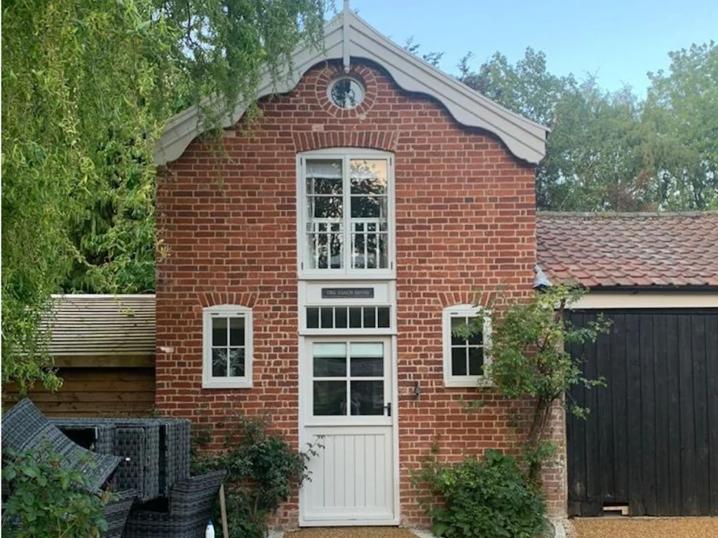 Red brick coach house with a scalloped white gable, a tall white-framed window above a stable-style door and a black timber garage door beside it
