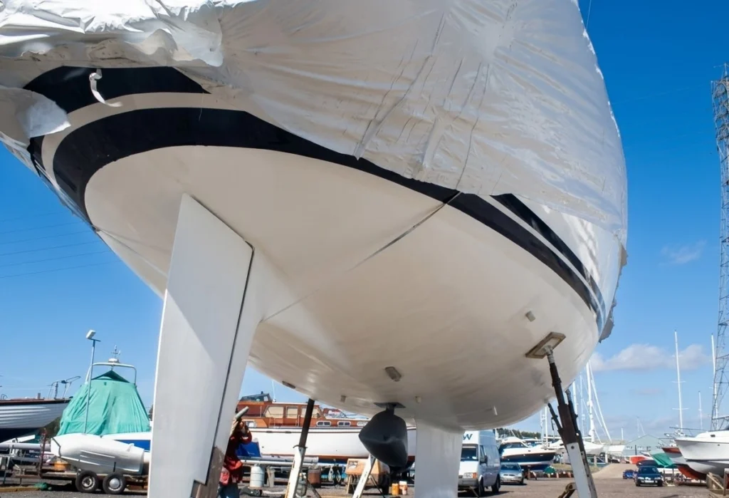 Yacht hull on support stands in a boatyard with a clean white underside, a black stripe and a protective cover over the deck