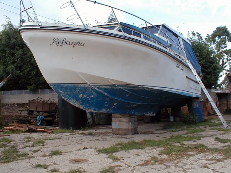 Motor cruiser named “Rebanna” on blocks in a yard, with a ladder against the side and old blue antifoul being removed from the lower hull