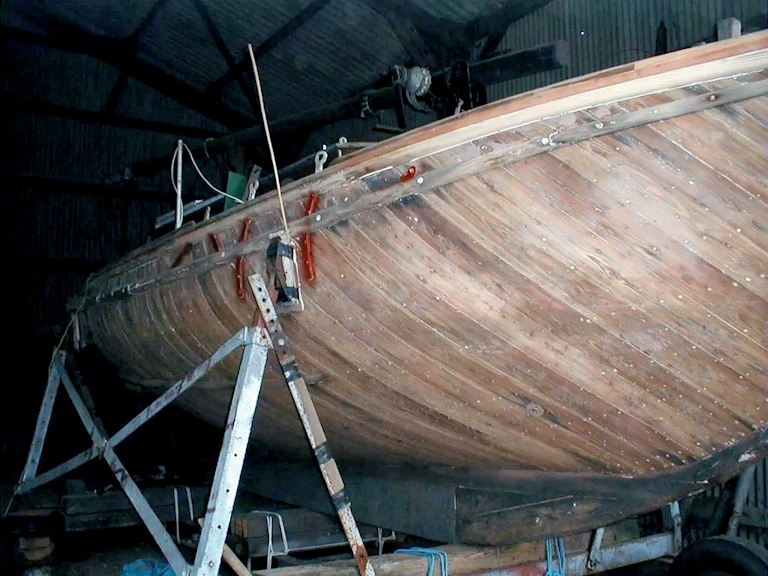 Wooden boat hull stripped back to bare timber planking, supported on a steel cradle inside a dark workshop