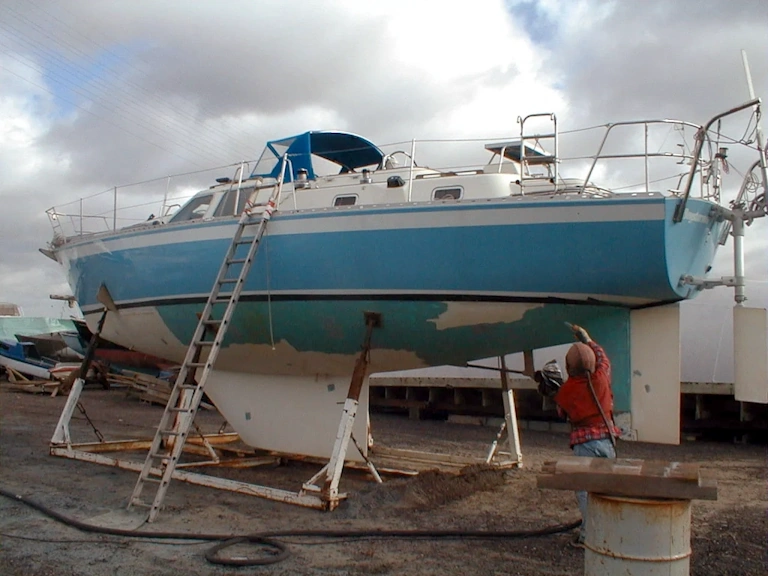 Blue and white sailing yacht on a cradle with a ladder against the hull as a worker in a red jacket strips paint from the lower hull
