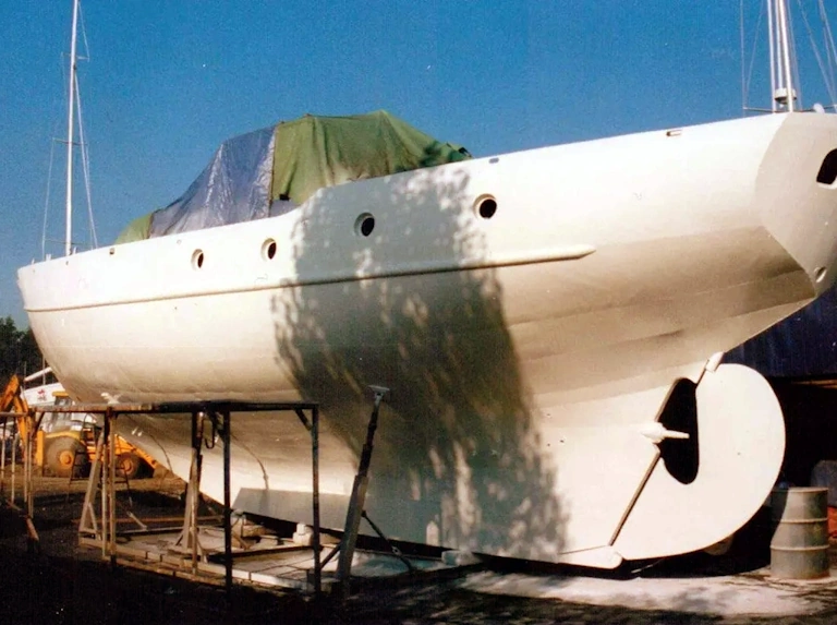 White yacht hull with round portholes resting on supports in a boatyard, with a tarpaulin over the deck and a tree shadow across the side