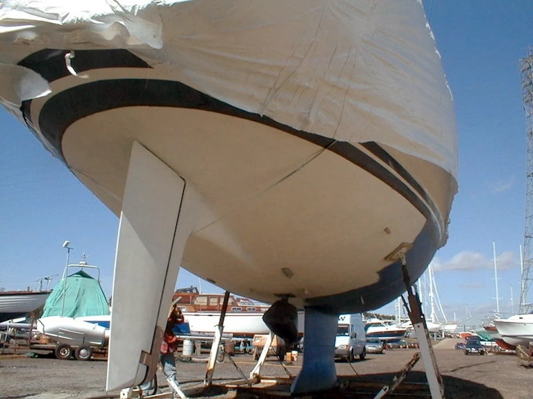 Sailing yacht out of the water on support stands in a boatyard, its hull stripped back to a pale surface with a person working beside the keel