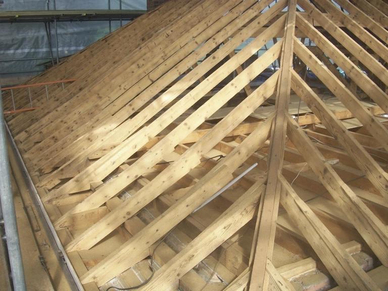 Timber roof structure with rows of pale rafters meeting at a central hip, photographed from above during construction with scaffolding at the side