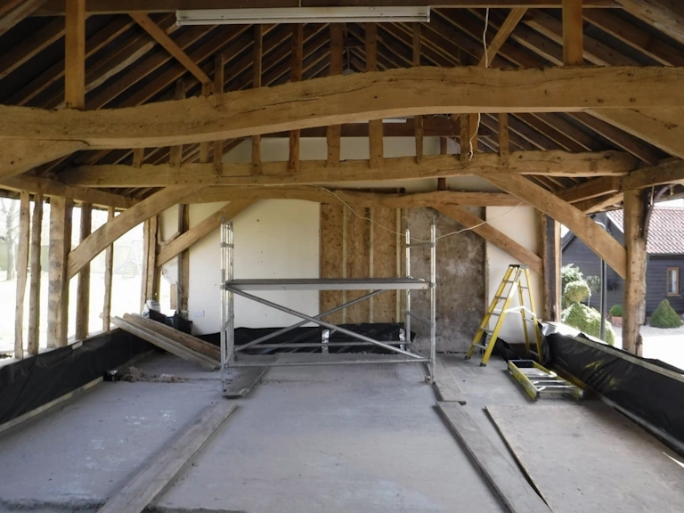 Open interior of an oak-framed barn with curved braces and tie beams overhead, a scaffold tower and stepladder on a bare concrete floor