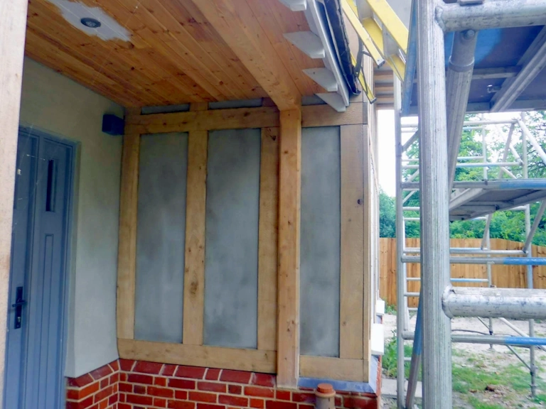 Oak-framed porch wall with grey infill panels above a red brick plinth, a pale blue door to the left and scaffolding to the right