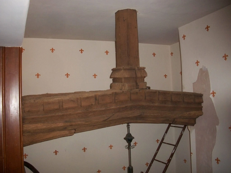 Large carved oak beam with a decorative dentil moulding and a square timber post above it, set against walls with a fleur-de-lis wallpaper pattern