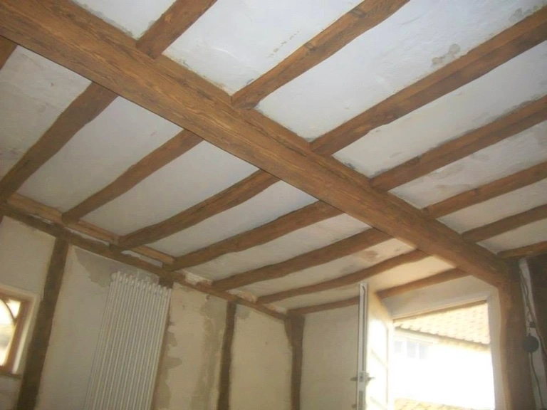 Exposed oak ceiling beams and joists in a room with white plastered panels between them, a radiator and a window with a view of tiled roofs