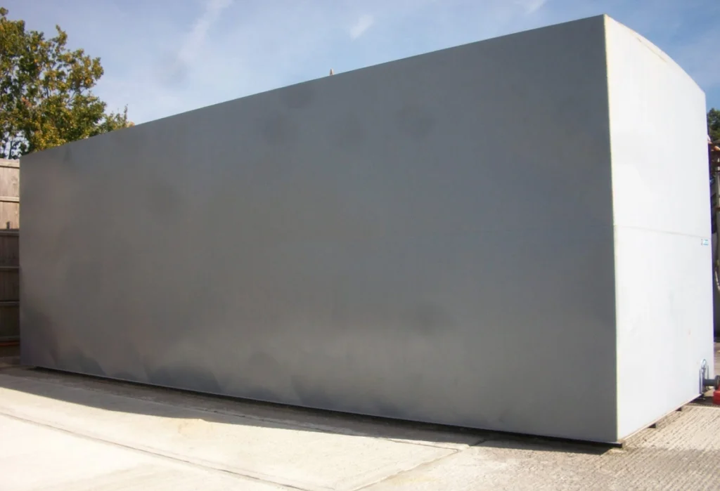 Large grey rectangular steel tank standing on a concrete yard, with trees and a timber fence behind