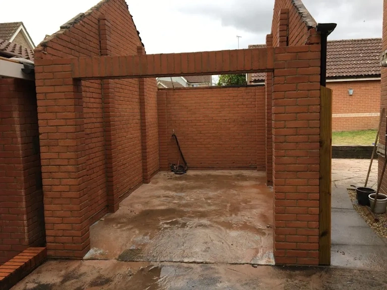 Newly built red brick garage structure with open front, brick lintel and a bare concrete floor, before roofing and doors were fitted