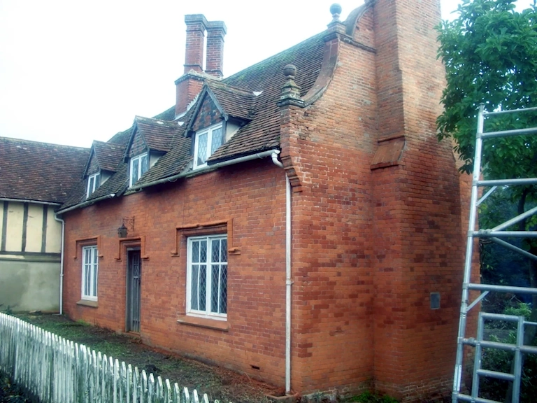 Aged red brick cottage with dormer windows, tall chimneys and leaded windows, with scaffolding to the right and a white picket fence in front