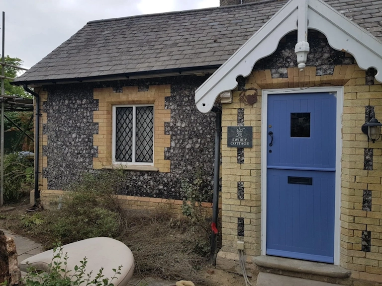 Single-storey flint cottage with yellow brick quoins, leaded windows, a white decorative gable and a blue stable door with a “Swirly Cottage” sign